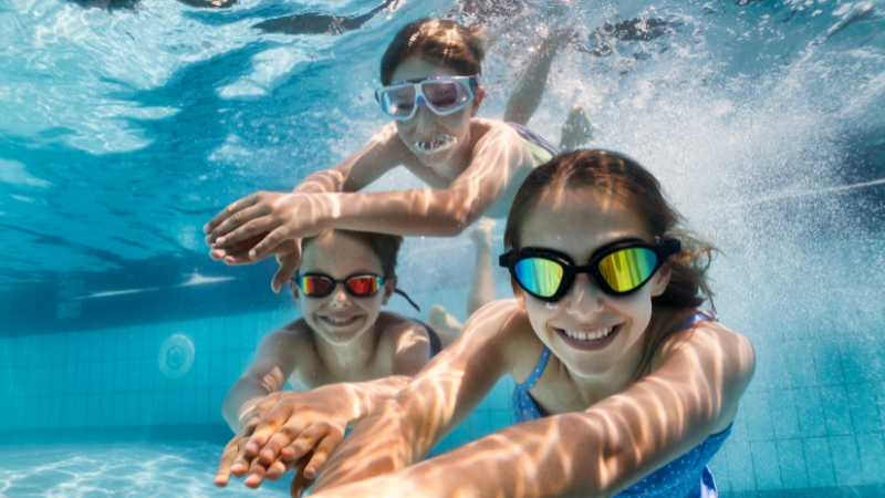 Children wearing googles underwater in a pool, swimming towards camera