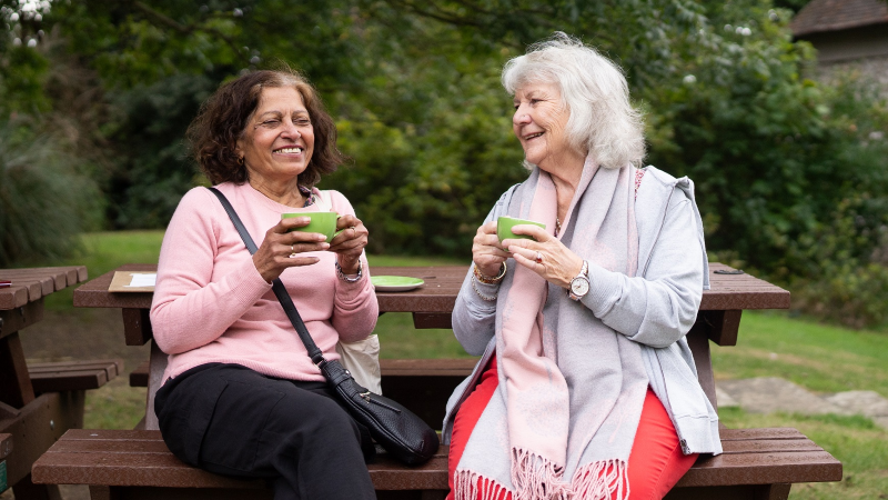 Two older ladies sat outside at a picnic bench drinking from cups and smiling