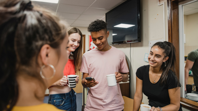 Group of young people in a youth club holding mugs and phones and chatting to each other happily
